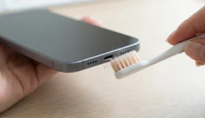 A close-up view of a person using a soft toothbrush to gently clean dust and debris from the bottom charging port and speaker grille of a smartphone to restore sound.