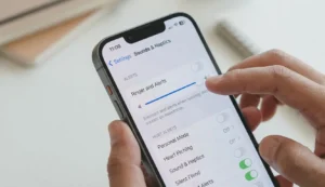 Close-up of a hand holding an iPhone, showing the Ring/Silent switch on the left set to 'Ring' (no orange), while the user simultaneously adjusts the "Ringer and Alerts" volume slider to maximum in the Settings menu (Sounds & Haptics).